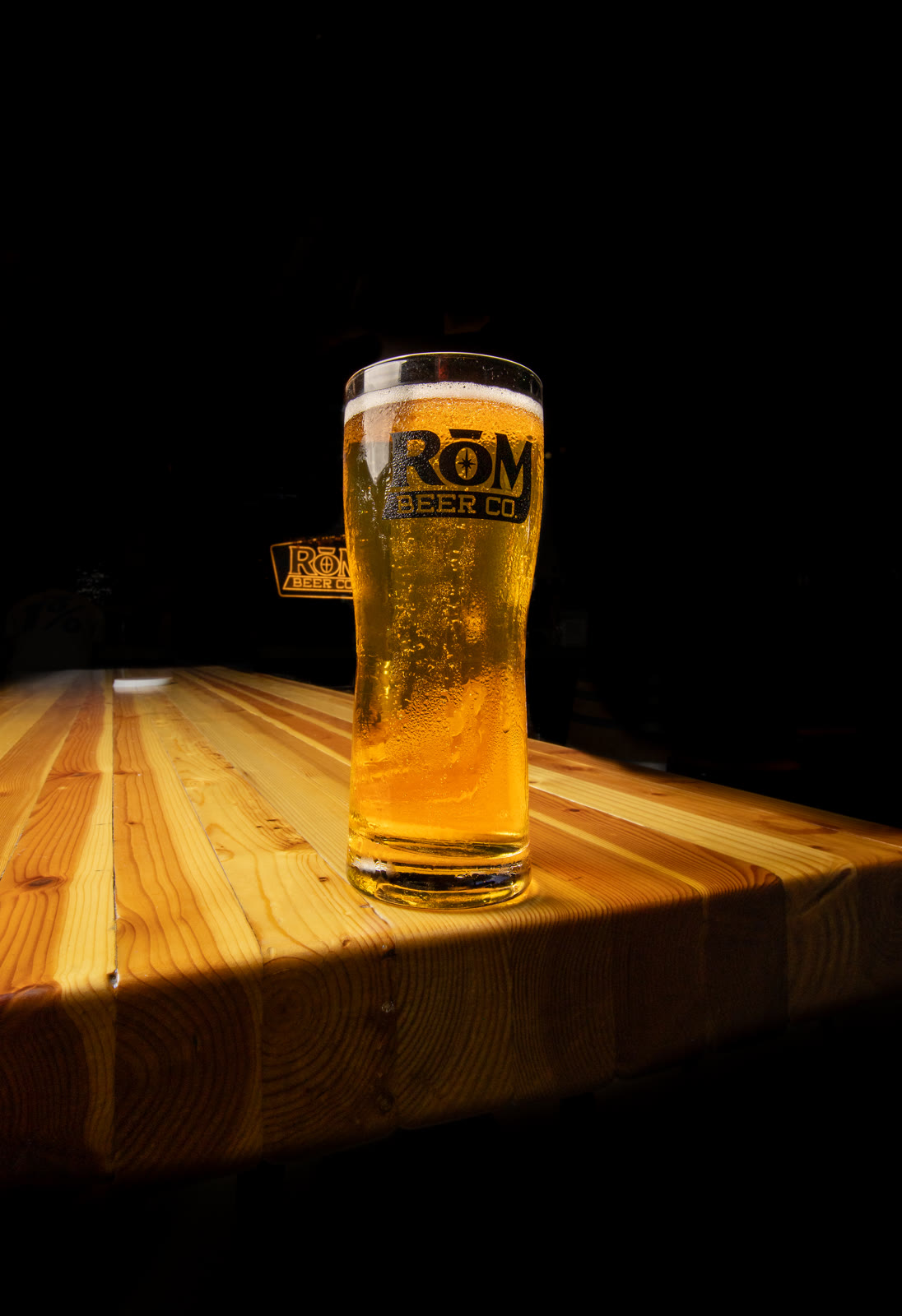 A glass of beer lit on the edge of a wooden table.